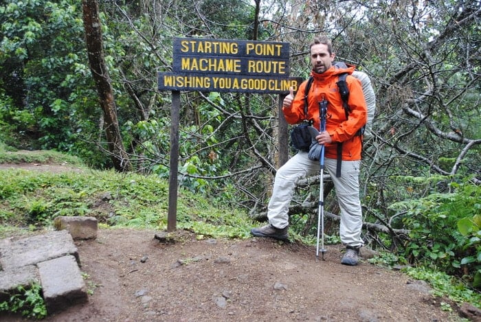 A male trekker in an orange jacket and grey pants stands next to a wooden sign in a lush forest that reads, "Starting Point Machame Route Wishing You A Good Climb."