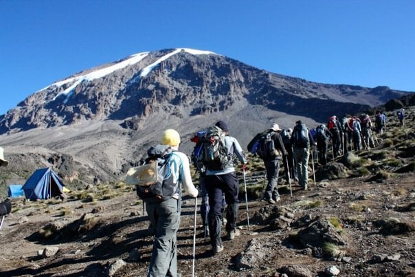 A long line of trekkers ascends a steep, rocky trail toward a large mountain peak with a distinctive white glacier at the top, passing a blue tent on the left.