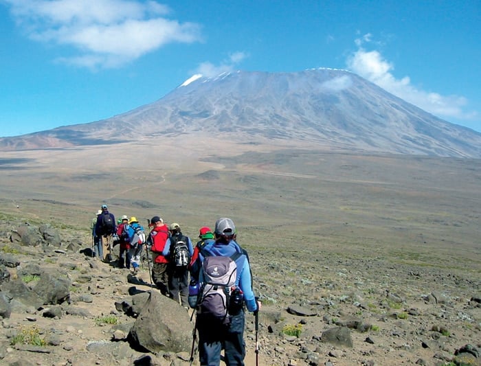 A group of trekkers with backpacks and hiking poles walks single-file along a rocky, barren trail toward a massive, broad-shouldered mountain with light snow on its summit.
