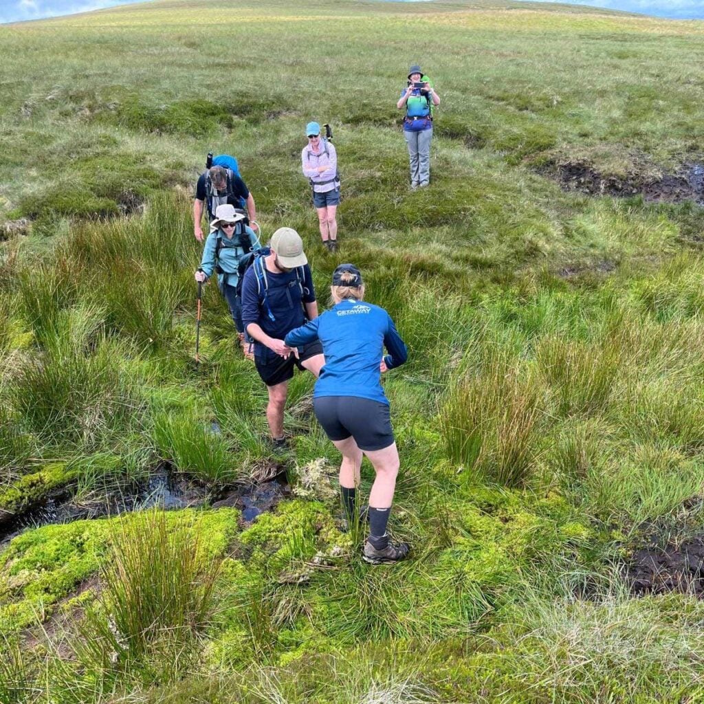 A group of six hikers maneuvers across a wet, marshy field of tall green grass and moss, with one hiker in the foreground reaching out to assist another over a small stream.