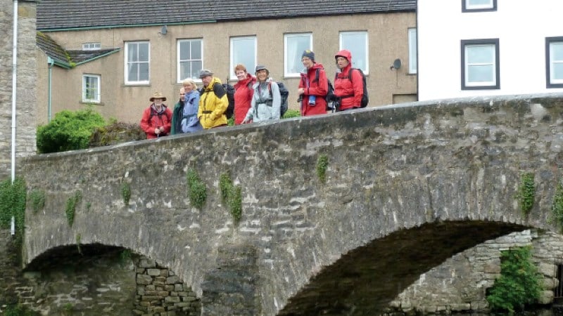 A group of seven hikers in colorful rain jackets stands along the edge of an ancient stone arch bridge, with traditional village buildings in the background.