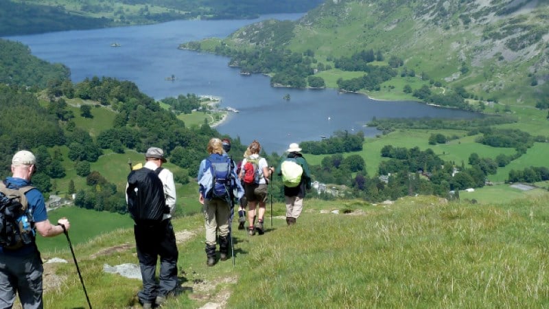 From a high vantage point, five hikers are seen from behind, descending a grassy mountain path. They are wearing hiking gear and large backpacks. In the valley below, a sprawling blue lake (likely in the Lake District) is nestled between rolling green hills and steep, rocky mountain slopes under a clear sky.