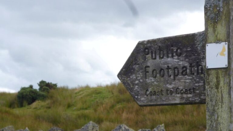 Wooden Public Footpath sign for the Coast to Coast walk in the English countryside, featuring rolling green hills and a cloudy horizon.