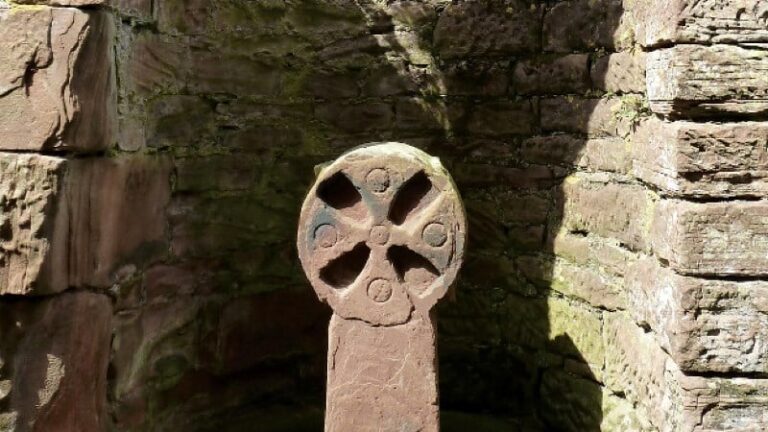 A close-up of an ancient, circular stone cross with four carved-out sections forming the cross shape. It is set against a background of a rugged, curved wall made of stacked brownish-red stones. Harsh sunlight creates deep shadows on the wall and the monument.