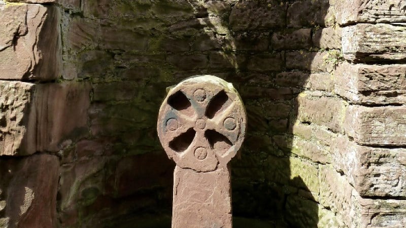 A close-up of an ancient, circular stone cross with four carved-out sections forming the cross shape. It is set against a background of a rugged, curved wall made of stacked brownish-red stones. Harsh sunlight creates deep shadows on the wall and the monument.