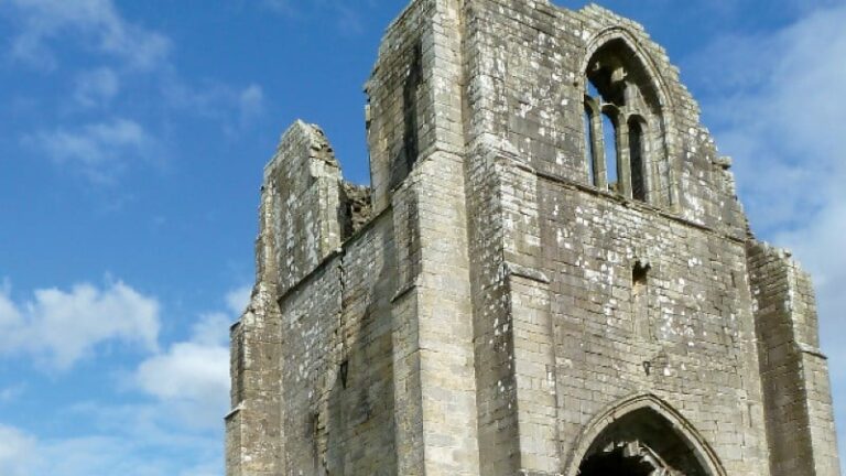 A low-angle shot of a massive, roofless stone structure made of grey blocks. The tower features arched window openings and a large Gothic-style pointed archway at the base, showing significant signs of age and weathering.