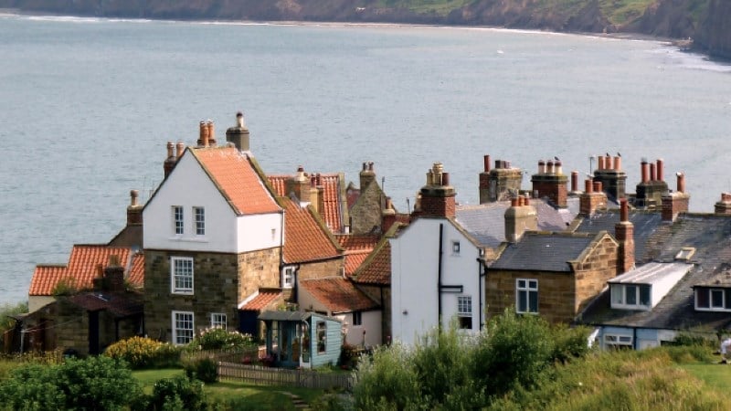 A high-angle shot of a seaside village. The houses feature traditional architecture with various chimney pots and small windows. A green grassy area with a small fence is in the foreground, and the shoreline curves into the distance under an overcast sky.