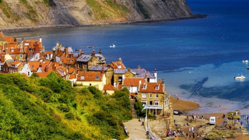A scenic overlook of a coastal town, likely Robin Hood's Bay. Traditional stone buildings with red-tiled roofs are nestled against a lush green slope. Below, a sandy beach meets the blue ocean where small boats are anchored. Massive, rugged cliffs rise in the background under a clear sky.