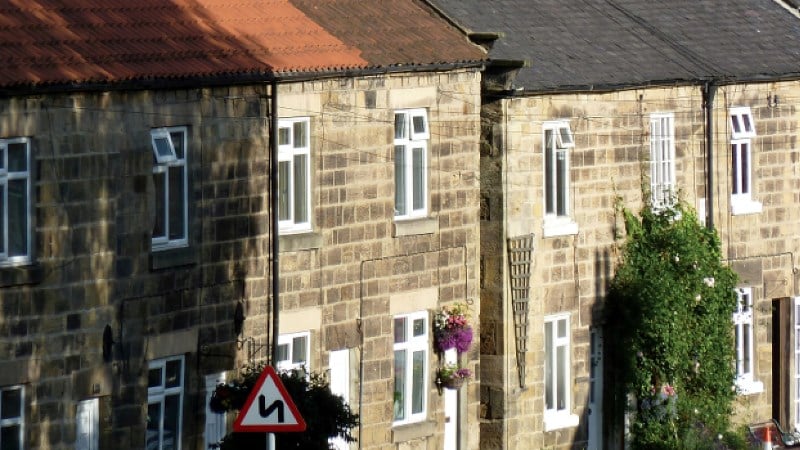 close-up view of stone-built terraced houses. The buildings feature light brown masonry, white window frames, and hanging flower baskets. A red-bordered triangular road sign with a black "winding road" symbol is visible in the foreground. Some greenery climbs the walls of the furthest house.