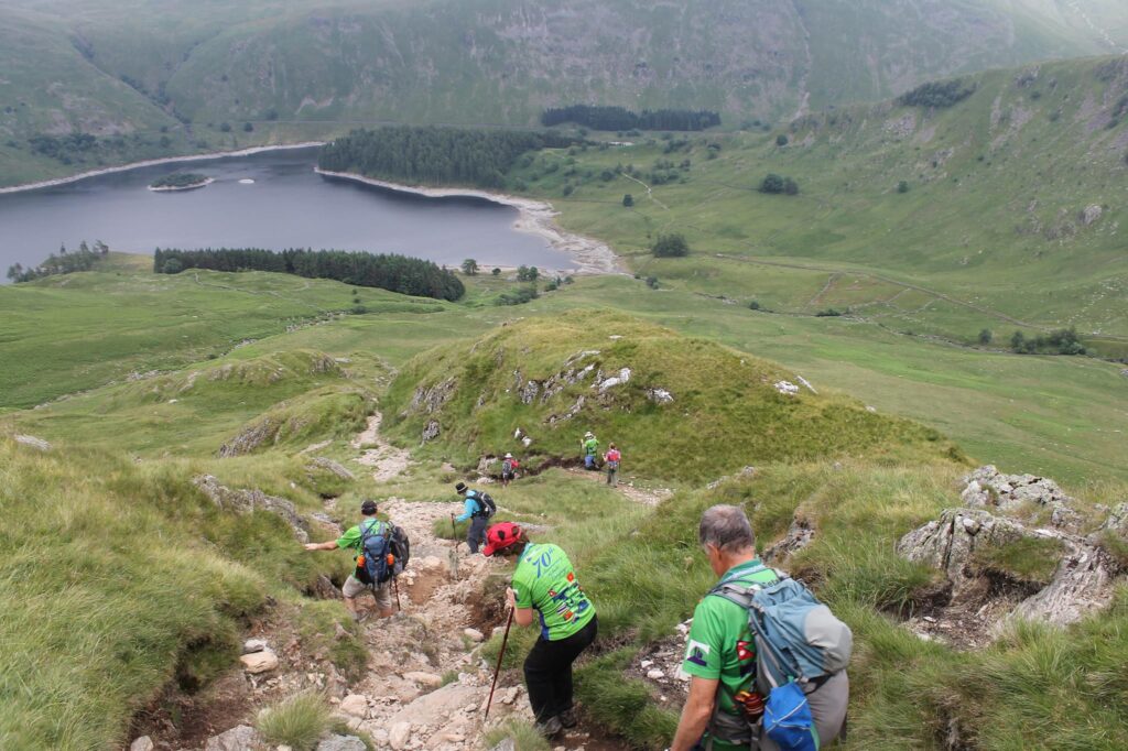 Hikers descending a rocky, grassy mountain path toward a large lake in a green valley.