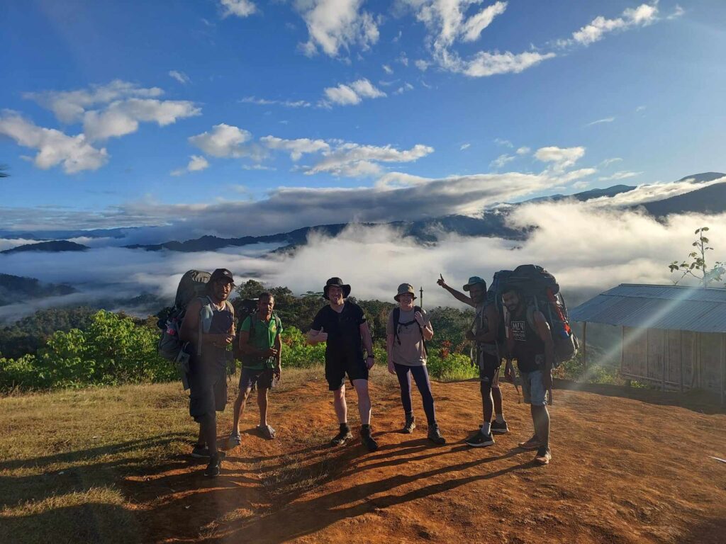 Group of six hikers standing on dirt clearing with backpacks; dressed in trekking gear; scenic mountainous landscape with lush green vegetation and dramatic mist settled in valleys behind; sky mostly clear with scattered clouds; small wooden structure with metal roof visible to right; scene conveys adventure, camaraderie, and natural beauty during hiking expedition.