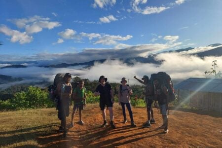 Group of six hikers standing on dirt clearing with backpacks; dressed in trekking gear; scenic mountainous landscape with lush green vegetation and dramatic mist settled in valleys behind; sky mostly clear with scattered clouds; small wooden structure with metal roof visible to right; scene conveys adventure, camaraderie, and natural beauty during hiking expedition.