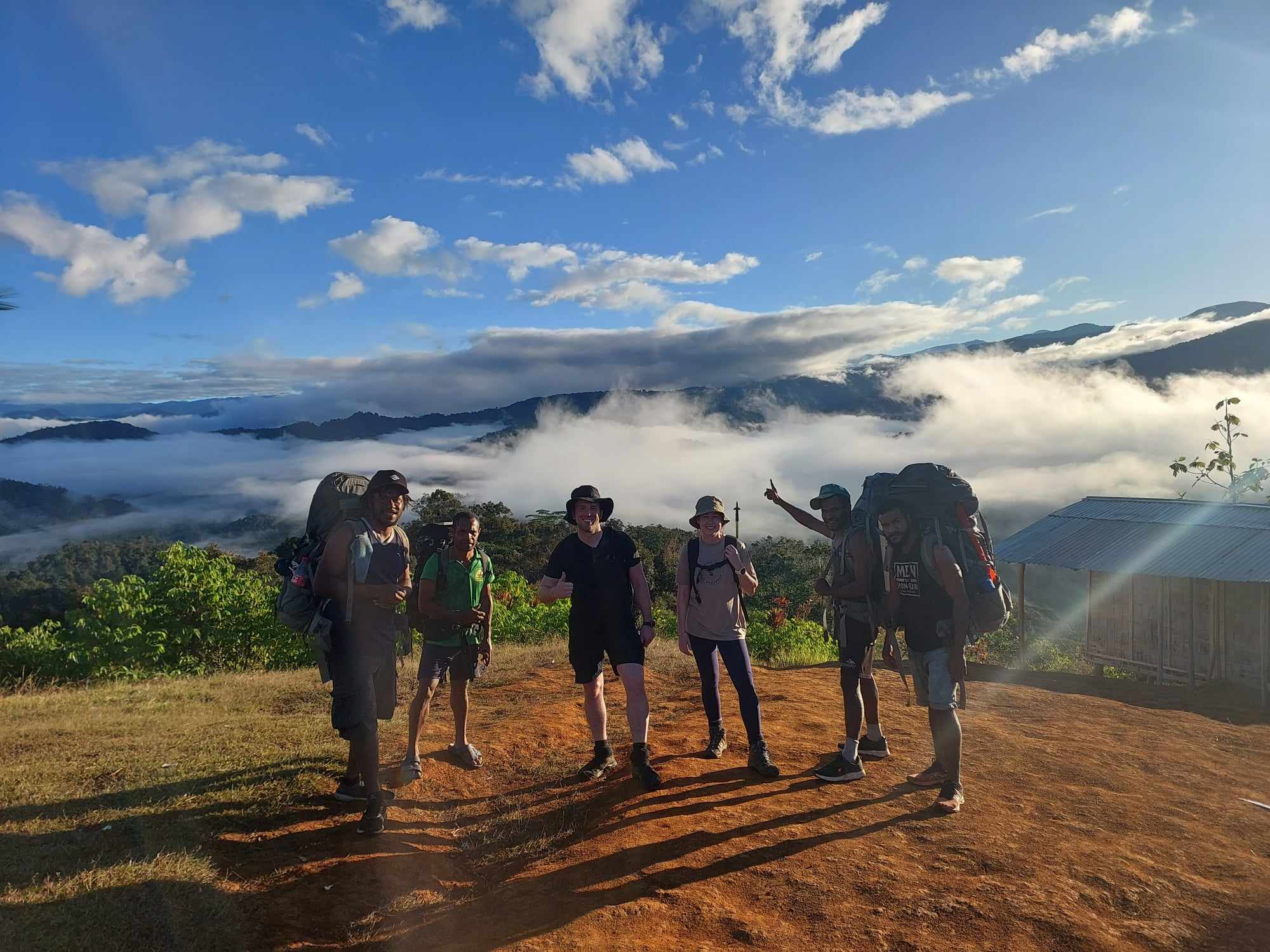 Group of six hikers standing on dirt clearing with backpacks; dressed in trekking gear; scenic mountainous landscape with lush green vegetation and dramatic mist settled in valleys behind; sky mostly clear with scattered clouds; small wooden structure with metal roof visible to right; scene conveys adventure, camaraderie, and natural beauty during hiking expedition.