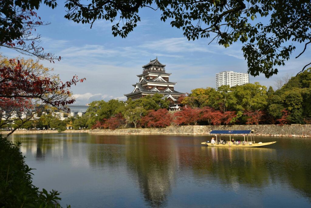 A traditional Japanese castle with multiple tiers and dark wooden walls, surrounded by colorful autumn trees and a calm moat with a sightseeing boat in the foreground.