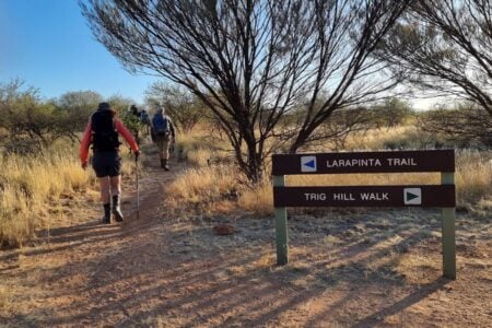 A group of hikers follows a dirt path into the scrubland, passing a brown wooden sign for the "LARAPINTA TRAIL" and "TRIG HILL WALK".