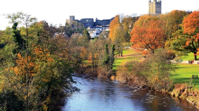 A wide scenic shot of a dark river. To the right, a lush green park features a bench and trees in peak fall colors. In the distance, the ruins of a stone castle and a tall square church tower rise above a small village